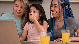 Zwei Frauen und ein Kind schauen gemeinsam auf einen Laptop und lachen.